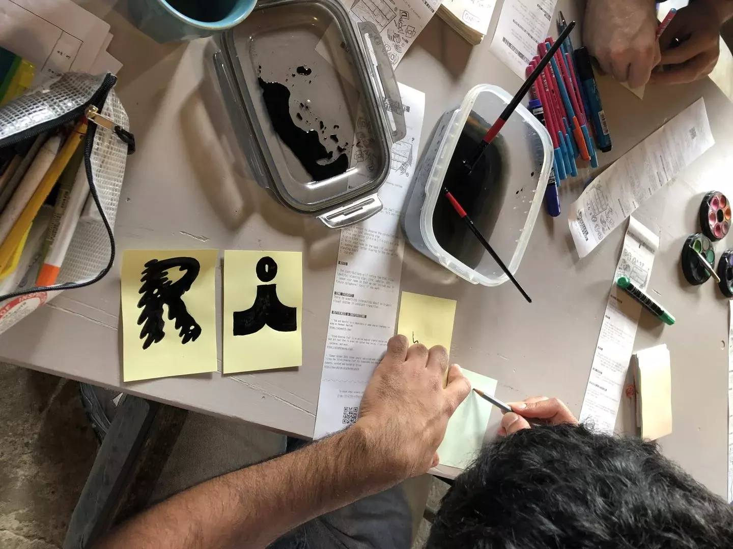 Detail of people working hands-on to draw some letters. There are two characters R and I drawn probably with some black ink, as suggested from the brushes and dark water bowl on the table.