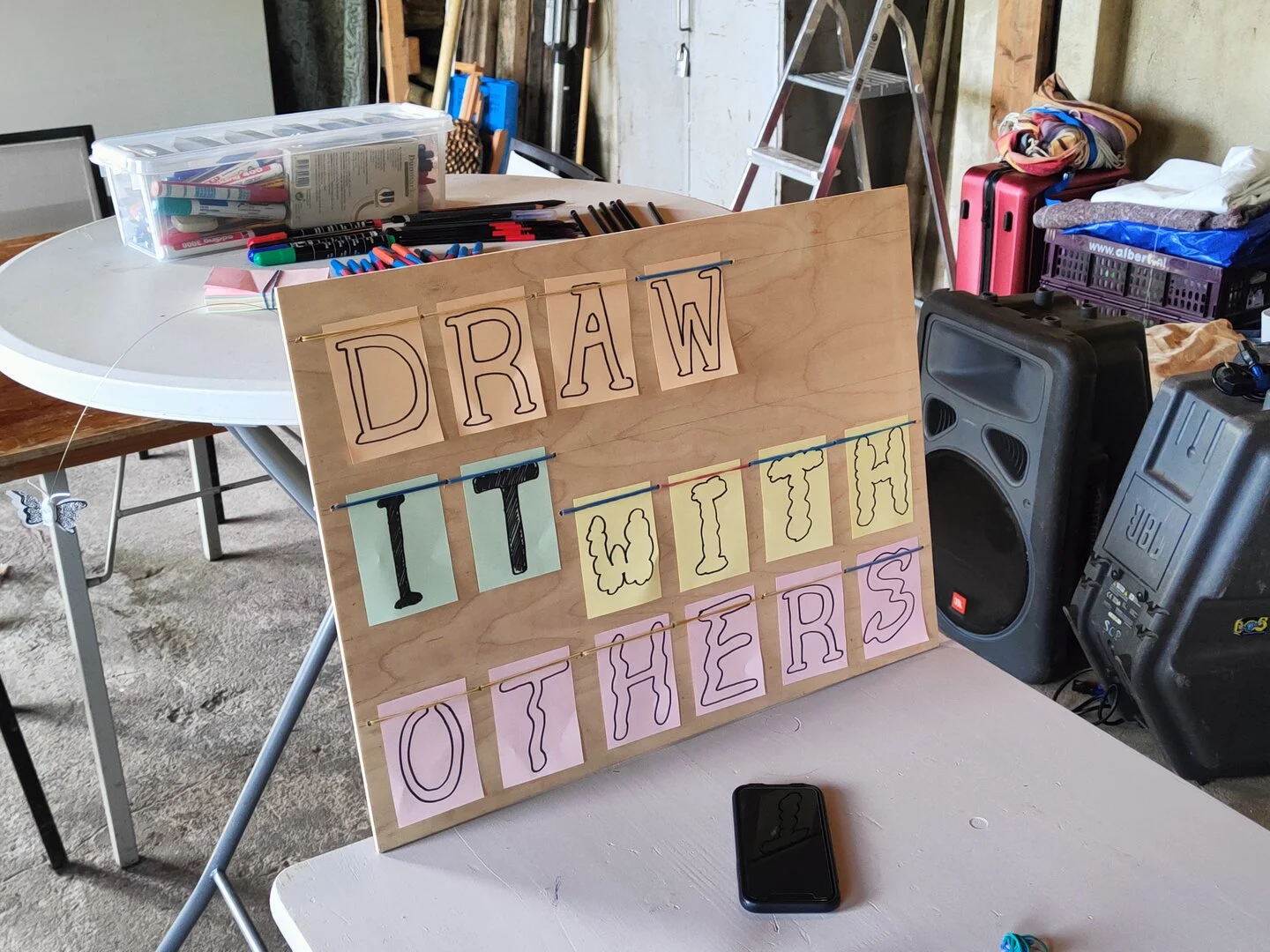 A wooden board in a messy workshop with some hanging cards forming the words: draw it with others. Each word is written with a definite style: some are more wobbly, some other more elegant.