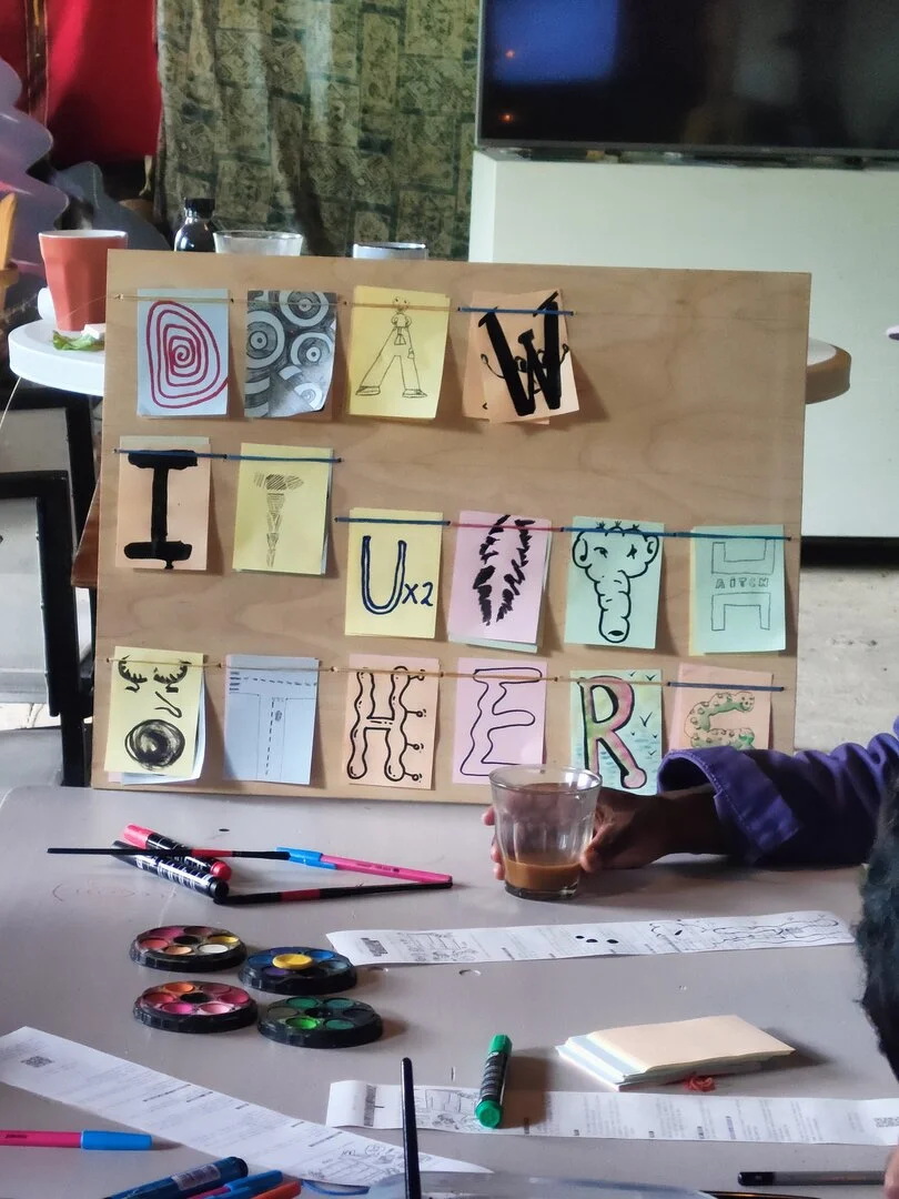 A wooden board with the words: draw it with others. Each letter is drawn in a different style: some of them are more comprehensible, other way more abstract. A "T" is made as the head of an elephant, an "O" is the face of someone howling, an "S" is a funny caterpillar, a "D" that is a spiral... 