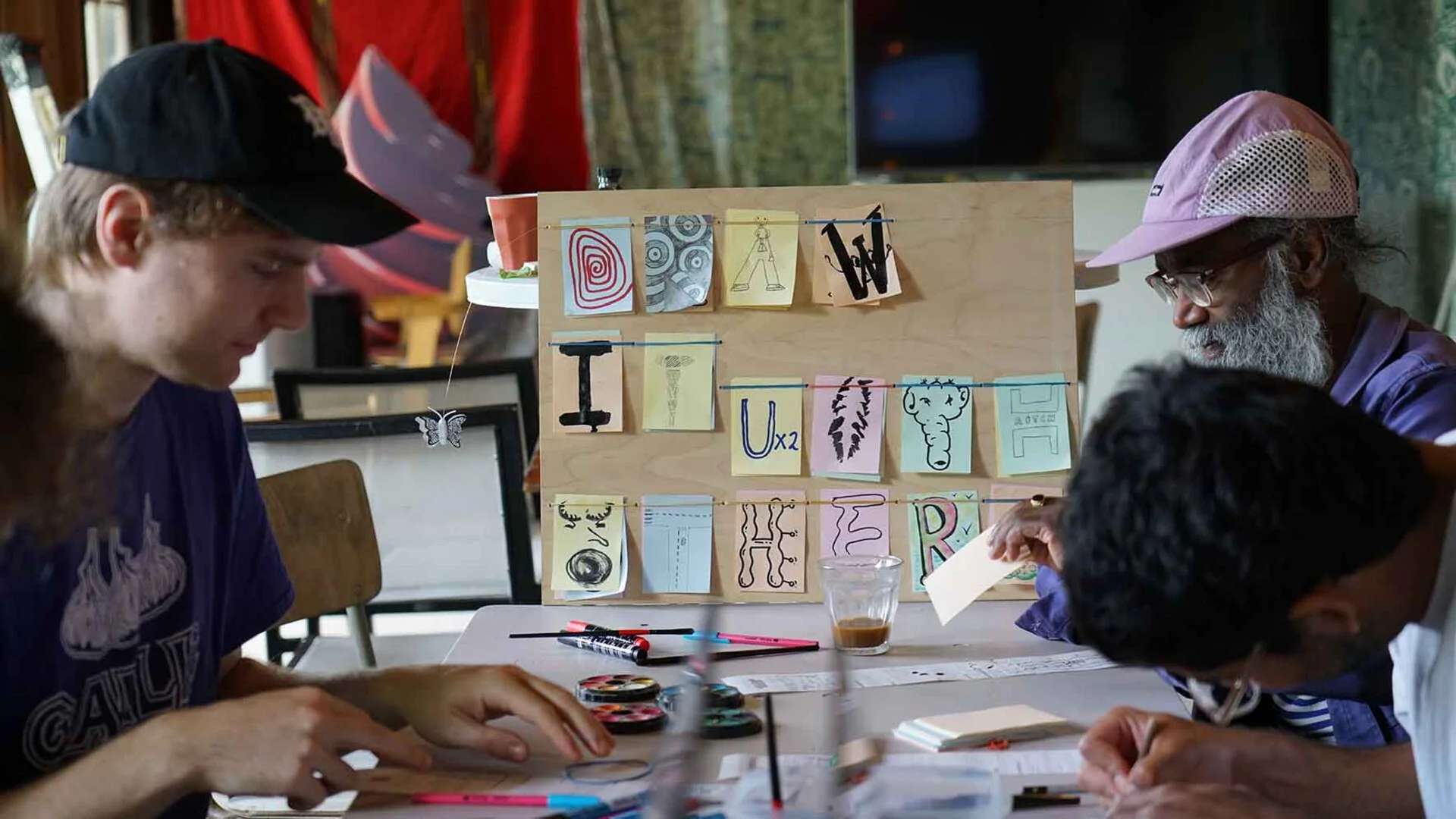 People busy around the table with the wooden board in the background. There is a shaky "E", a "W" that is literally U times 2, a "T" drawn as a crossroad, and a wonky "H" with some tentacles on one side that reminds of DNA.