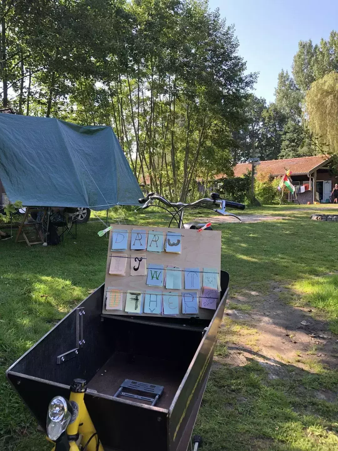 The wooden board in a cargo bike parked in a sunny camping site. There's a willow and some other trees in the background.