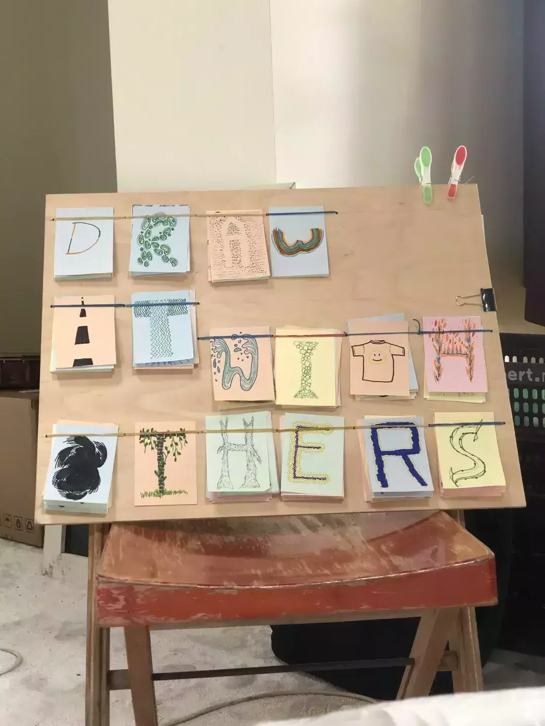 Overall picture of the wooden board with lot of different characters. A "W" is a double rainbow upside-down, a t-shirt as a "T", some heavy brush strokes make for an obscure "O", some dashed centerlines on a road form an 'i'...